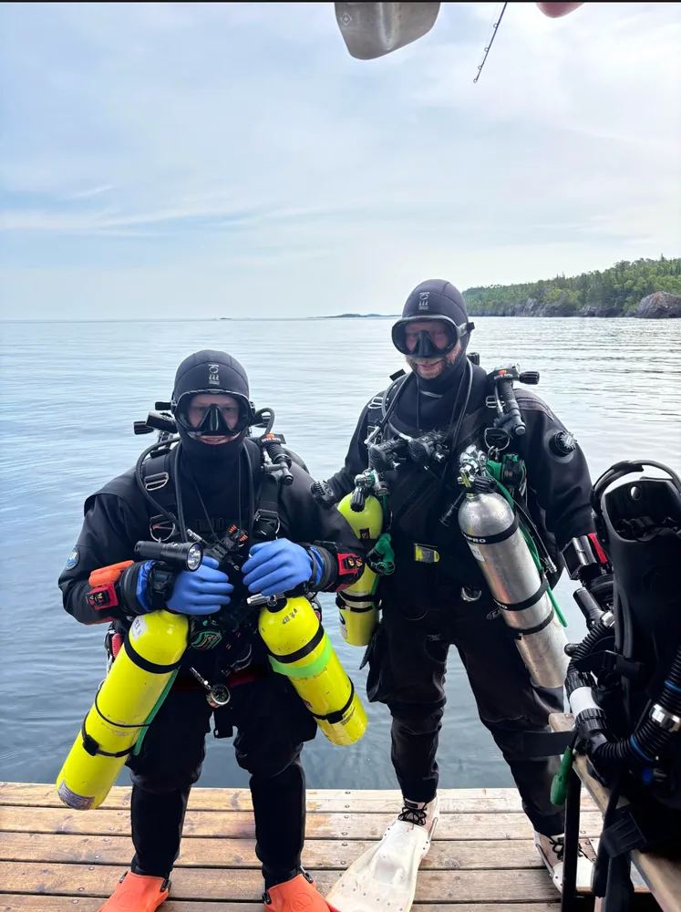 Two divers in drysuits and dive gear on a dock before a cold-water dive